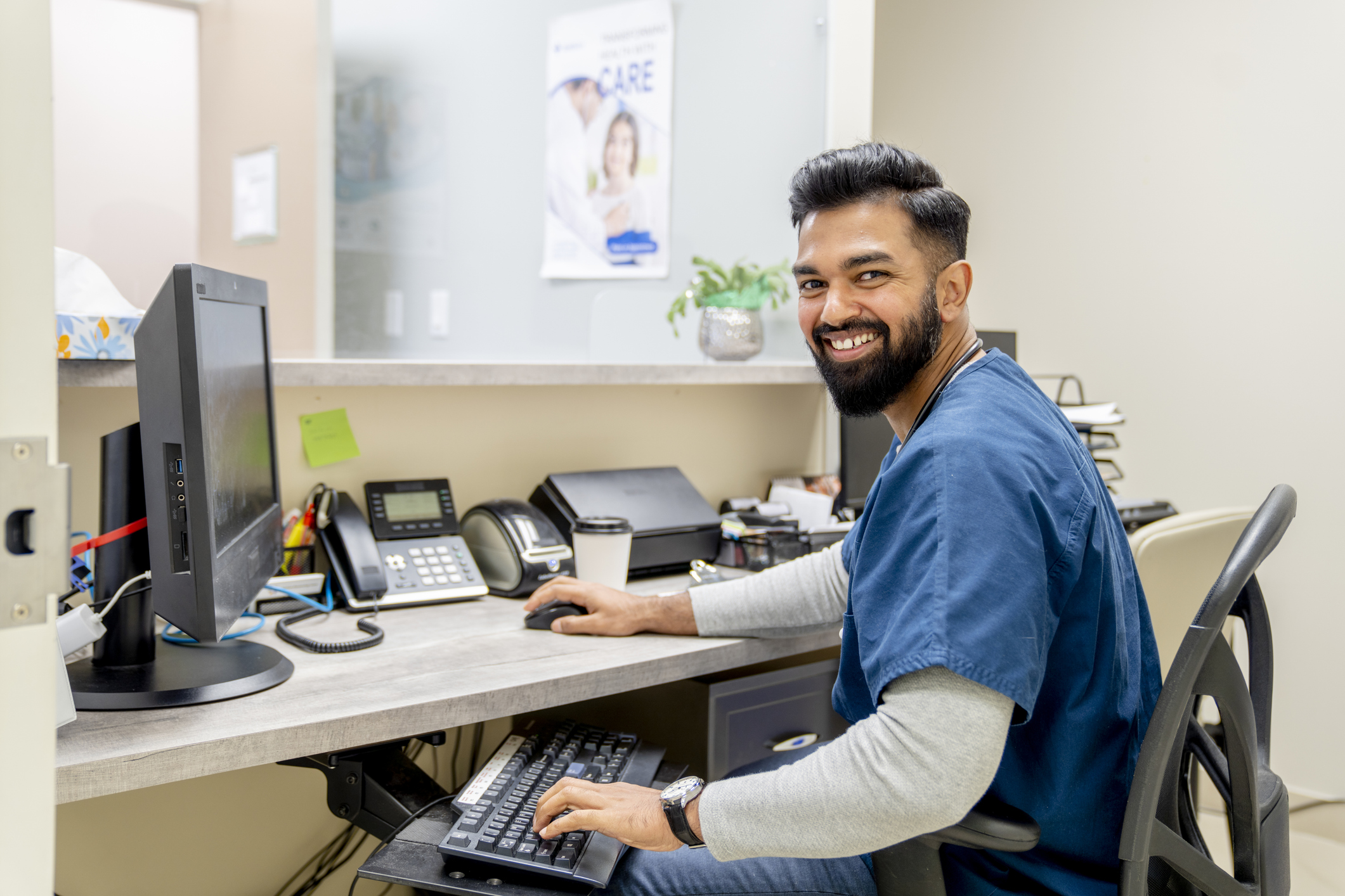 A handsome young male nurse of Indian descent is standing at the front counter of a medical clinic. He has a computer at his desk. He is wearing blue medical scrubs. He is looking at the camera and smiling widely.